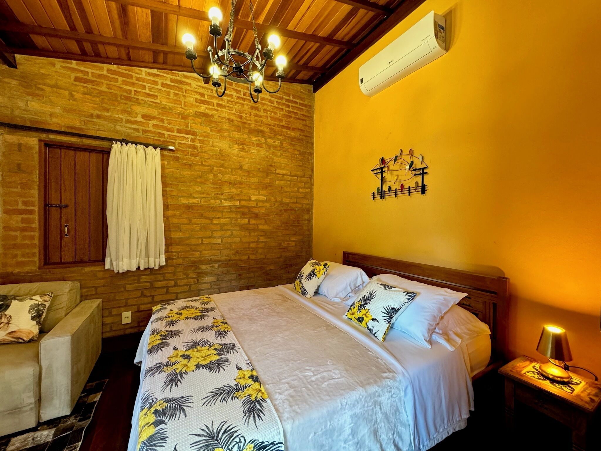Um quarto com parede de tijolos, teto de madeira, cama com roupa de cama floral, mesa de cabeceira de madeira com abajur e ar-condicionado fixado na parede.
