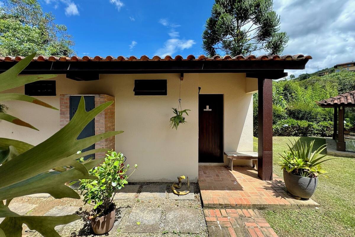 Una pequeña casa con techo de tejas, entrada de ladrillo y un porche pequeño. Hay plantas en macetas en frente de la casa y un árbol grande al fondo.