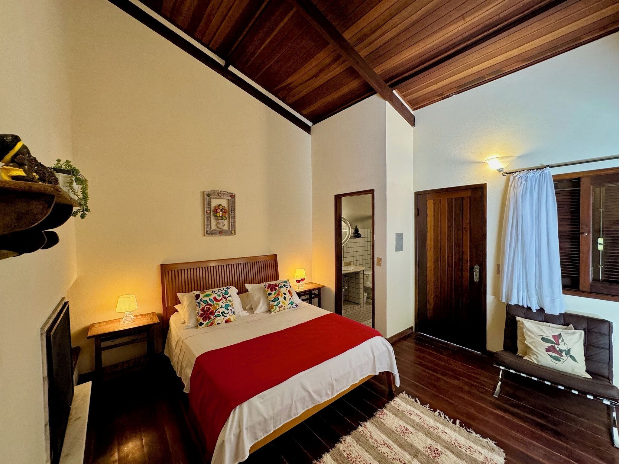 A bedroom with a wooden ceiling, a bed with a red and white bedspread, a bedside table with a lamp, a mirror, and a window with curtains.