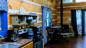 A rustic cabin interior with wooden walls, a kitchen area with a stove, sink, and cabinets, and a dining table with chairs.