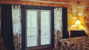 A log cabin bedroom with a large window, wooden ceiling beams, and a bed with a quilt.