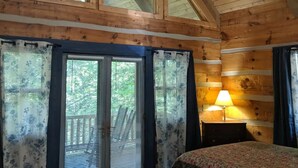A cabin with a high ceiling, wooden beams, a bed with a floral bedspread, a ceiling fan, and a window with blinds.