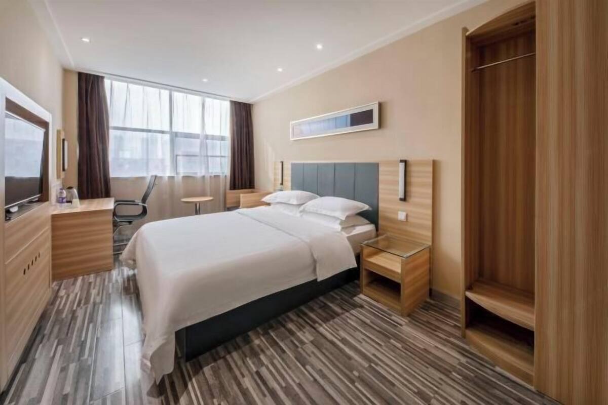 Ein modernes Hotelzimmer mit einem großen Bett, einem Holzschrank, einem Schreibtisch und einem großen Fenster mit Vorhängen.