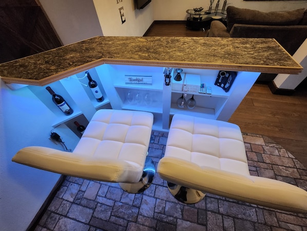 A bar area with a marble countertop, two cushioned seating, wine bottles, glasses, and a sign that reads "Our Life Our Story Our Home".