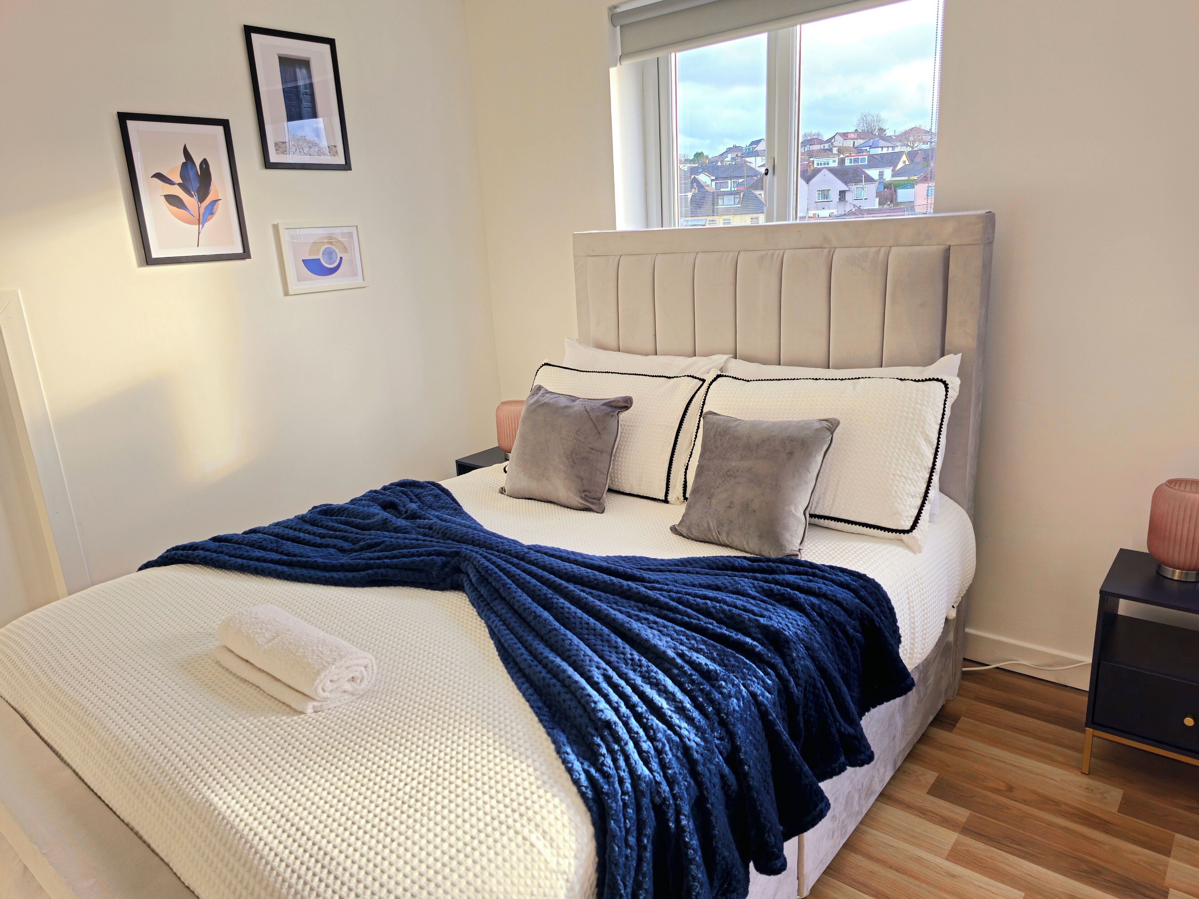 A bedroom with a large bed, a window with a view of houses, and framed pictures on the wall.