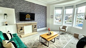A modern living room with a fireplace, a sofa, a coffee table, and a television displaying the AirUs logo. - Spacious Haven with Free Parking (Glasgow)