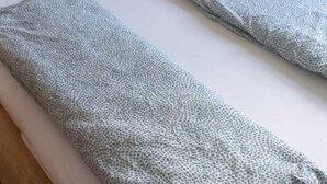 Una cama con colchón blanco y dos almohadas, cabecera de madera y una mesita de noche con superficie negra.