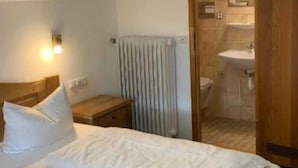 Una habitación de hotel con una cama grande, cabecera de madera, una mesita de noche y un radiador. Se ve un baño a través de una puerta abierta.