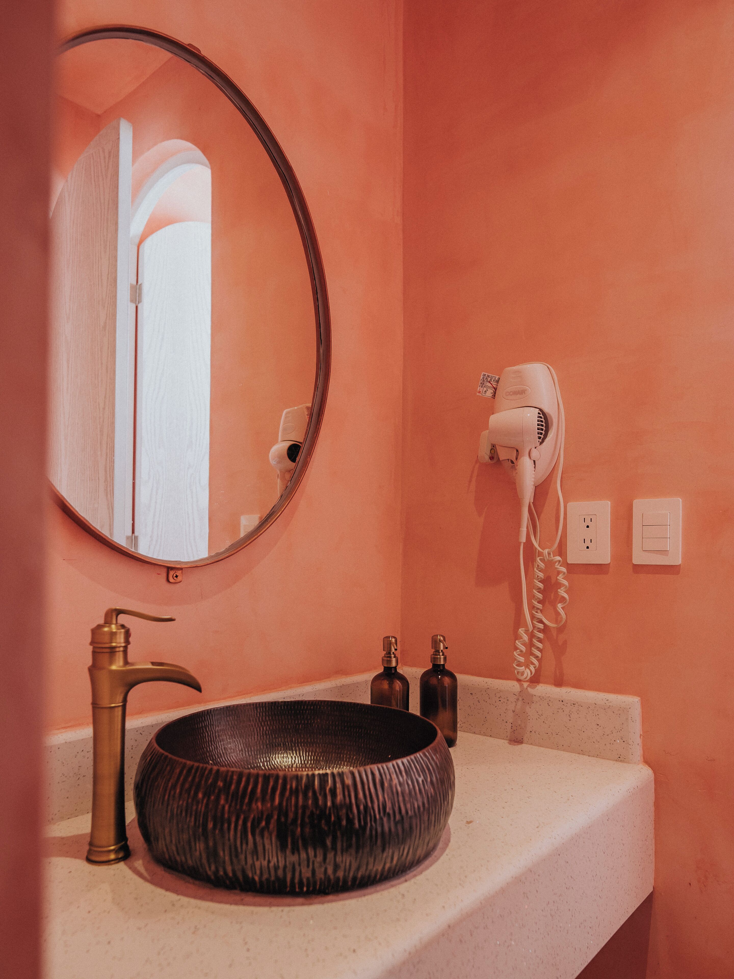 Une salle de bain avec un miroir rond, un robinet doré, un lavabo à texture, un sèche-cheveux et deux petits flacons posés sur le comptoir.