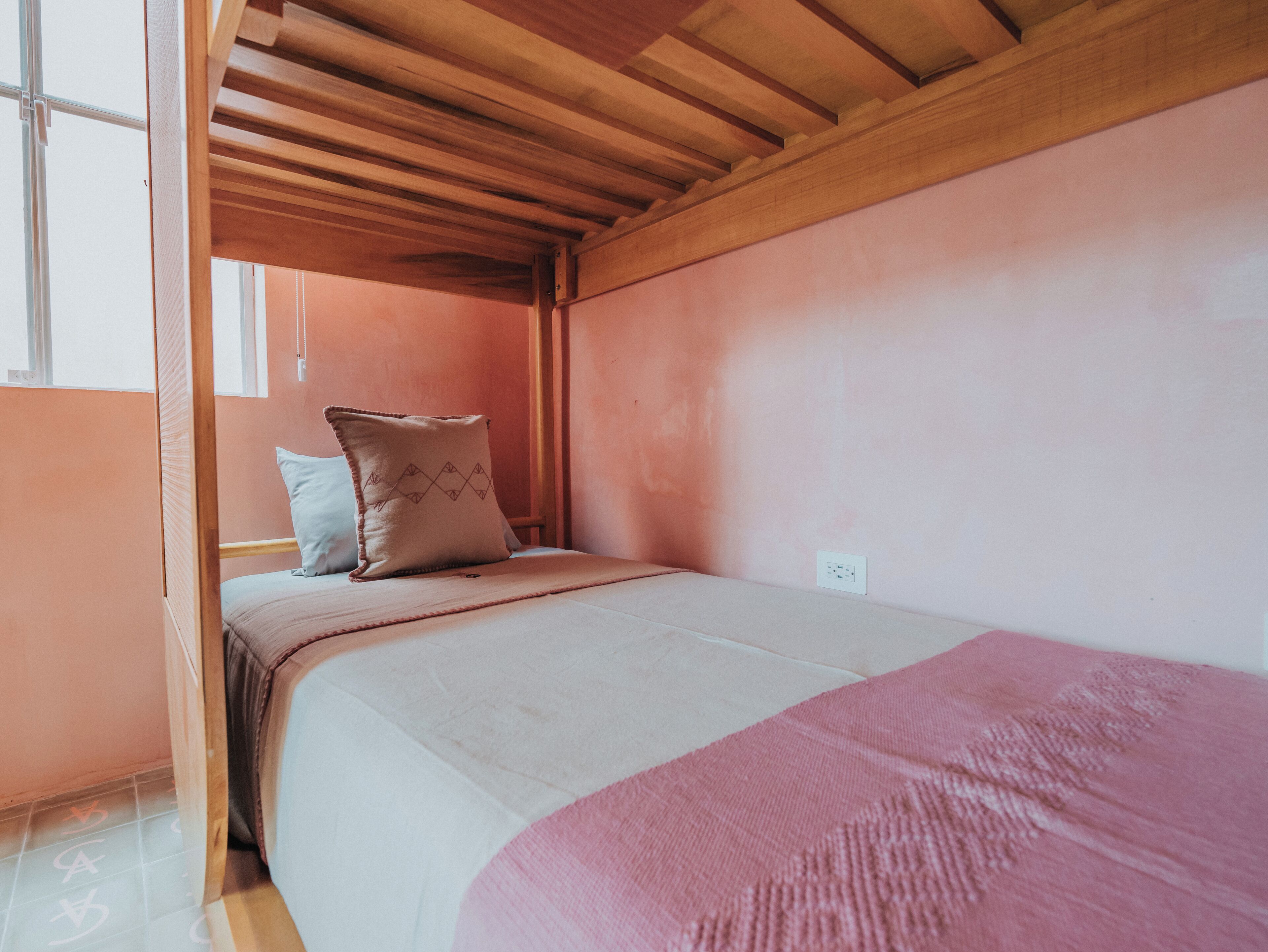 A bunk bed room with a wooden ceiling, a pink wall, and a window.