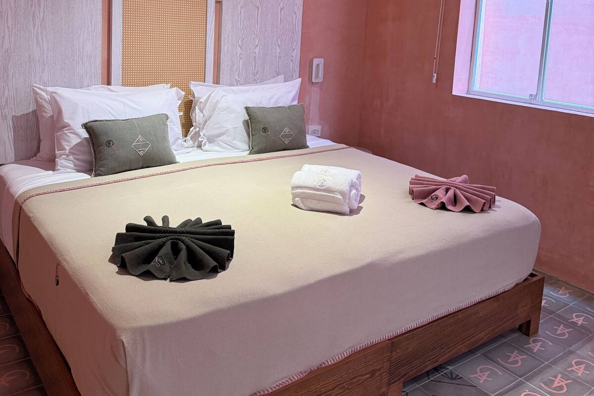 A neatly made bed with pillows and a folded towel, a wooden headboard, a window with blinds, and patterned floor tiles.