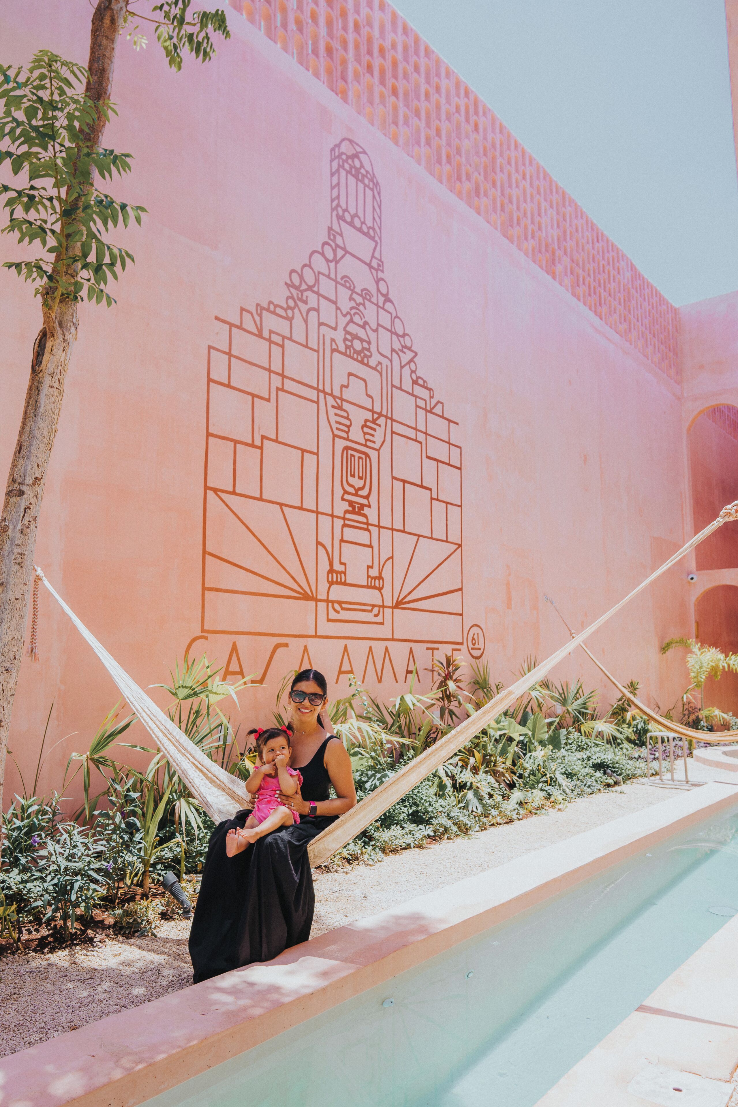 A woman in a black dress sitting on a hammock with a child, in front of a wall with a geometric design and the text "CASA AMATE".