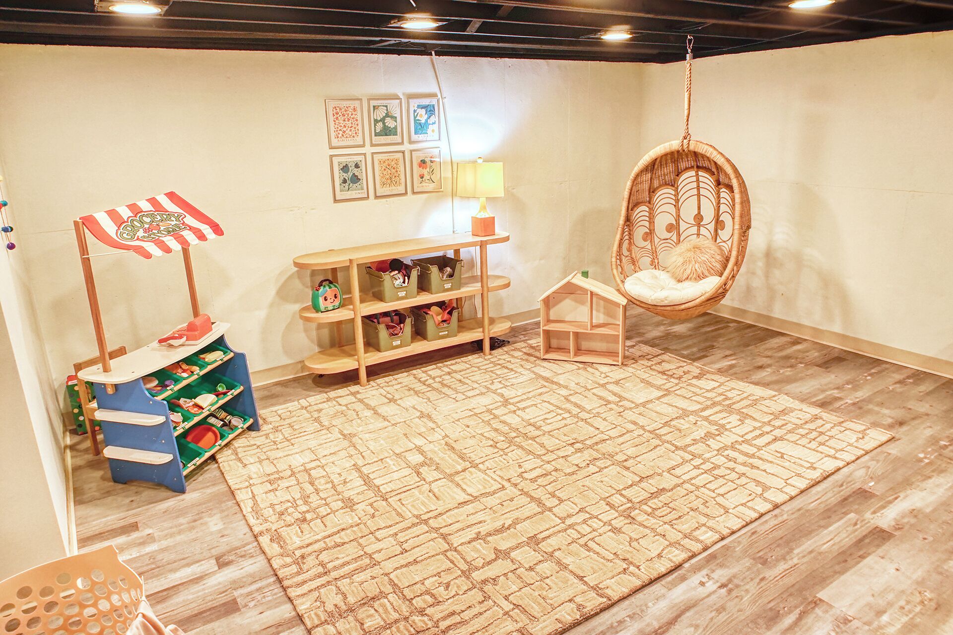 A cozy room with a hanging chair, a small table with a lamp, a toy kitchen set, and a patterned rug.