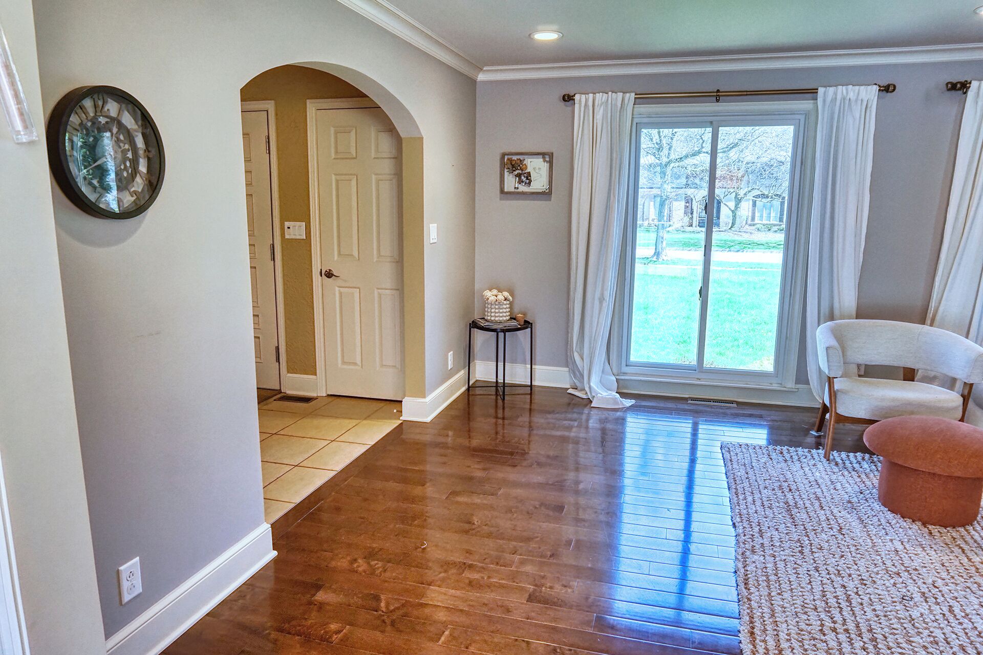 A spacious room with hardwood flooring, a white armchair, a small side table, and a large window with curtains.
