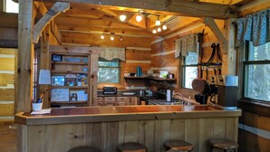A rustic cabin interior with a wooden bar area, bar stools, a kitchen with a stove, and a window with curtains.