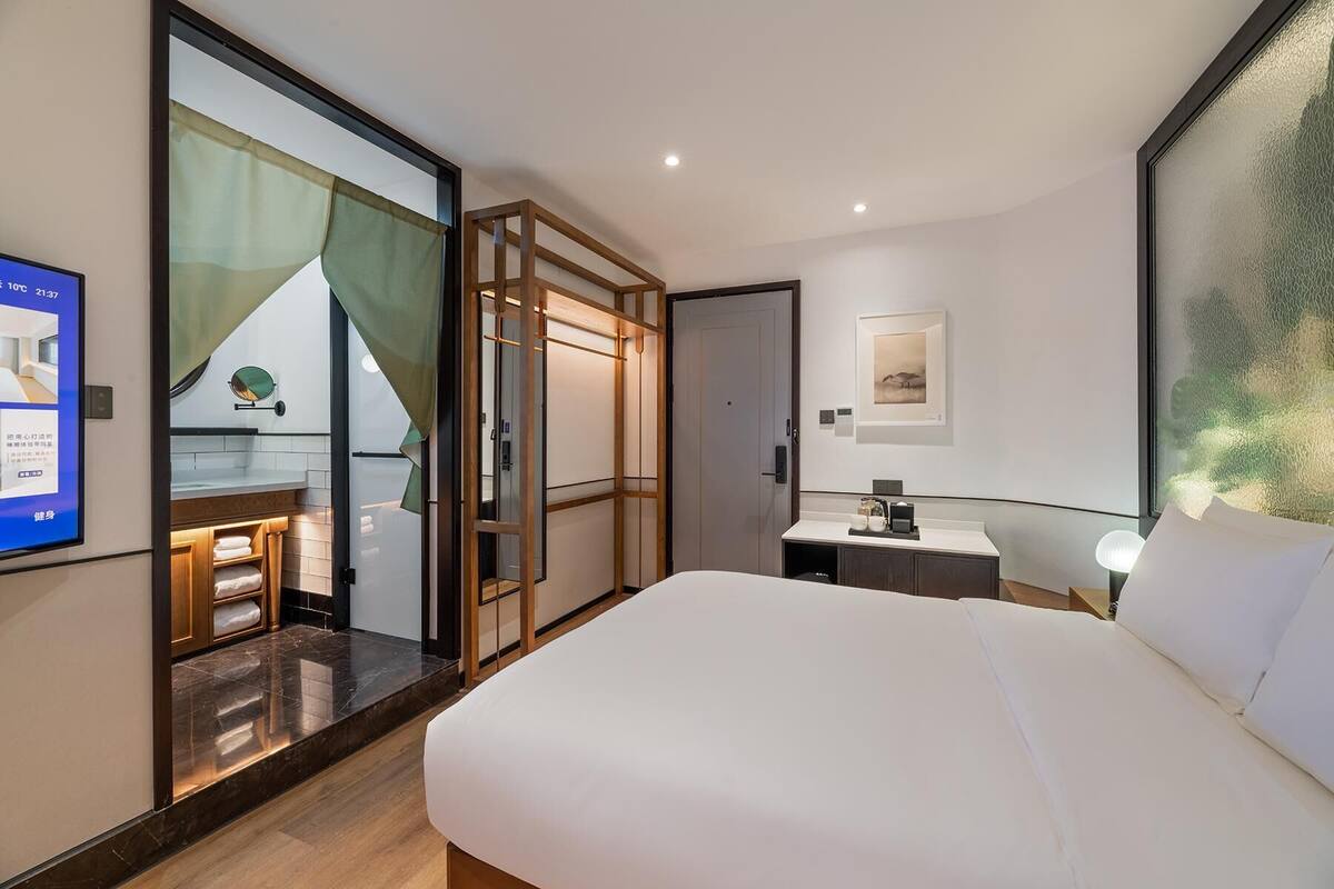 A hotel room with a large bed, a small desk with a lamp, a bathroom with a mirror and towel rack, and a sliding glass door leading to a balcony.