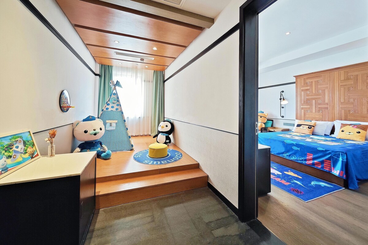 A hotel room with a wooden staircase, a bed with a blue ocean-themed bedspread, a wooden headboard, and a small play area with a tent and stuffed animals.