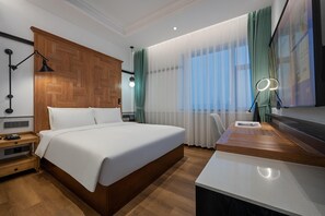 Habitación de hotel moderna con cabecera de madera, una cama grande, un escritorio con lámpara y vistas de la ciudad a través de cortinas translúcidas.