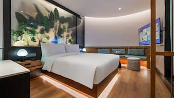 Quarto moderno com cama grande, mesa de cabeceira com abajur, um espaço de estar com um banco e uma TV fixada na parede.