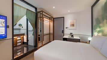 Una habitación de hotel con una cama grande, un escritorio pequeño con lámpara, un baño con espejo y perchero, y un ventanal corredero que da a un balcón.