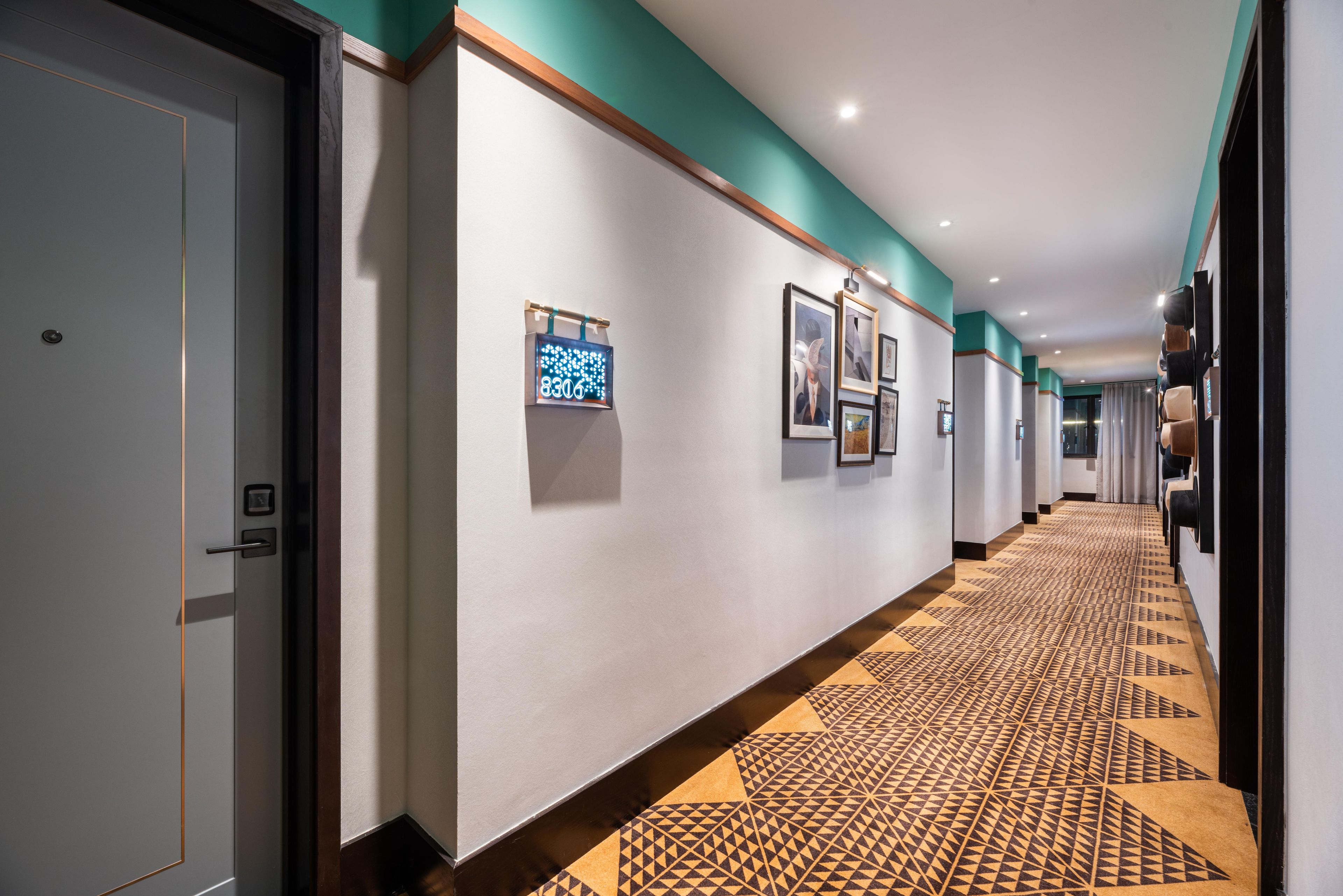 A hotel hallway with a patterned carpet, framed pictures on the walls, and a door with a digital display.