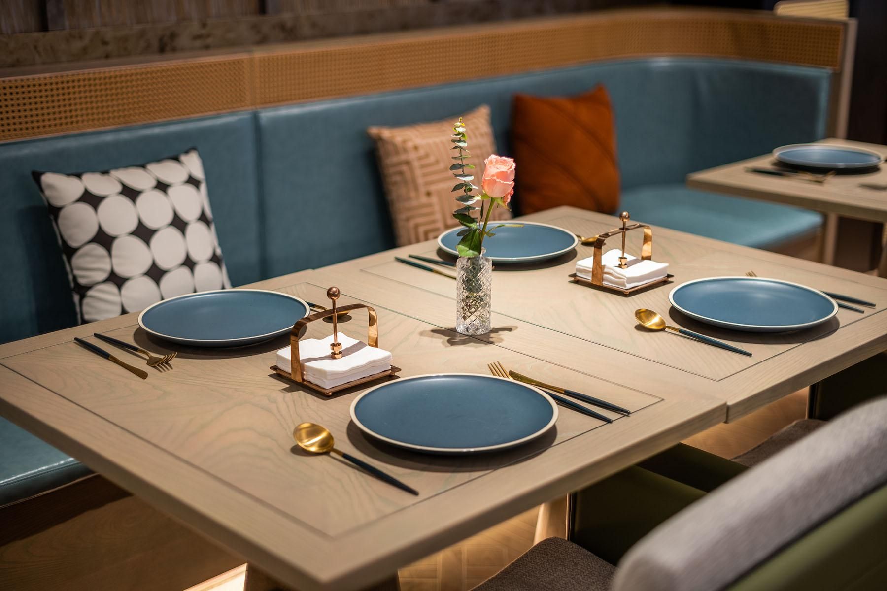 A dining table set with blue plates, gold cutlery, and a vase with a rose.