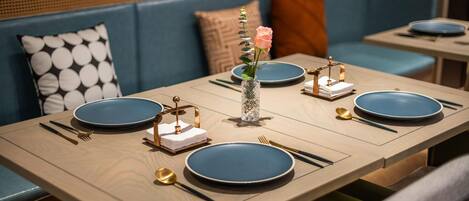 A dining table set with blue plates, gold cutlery, and a vase with a rose.