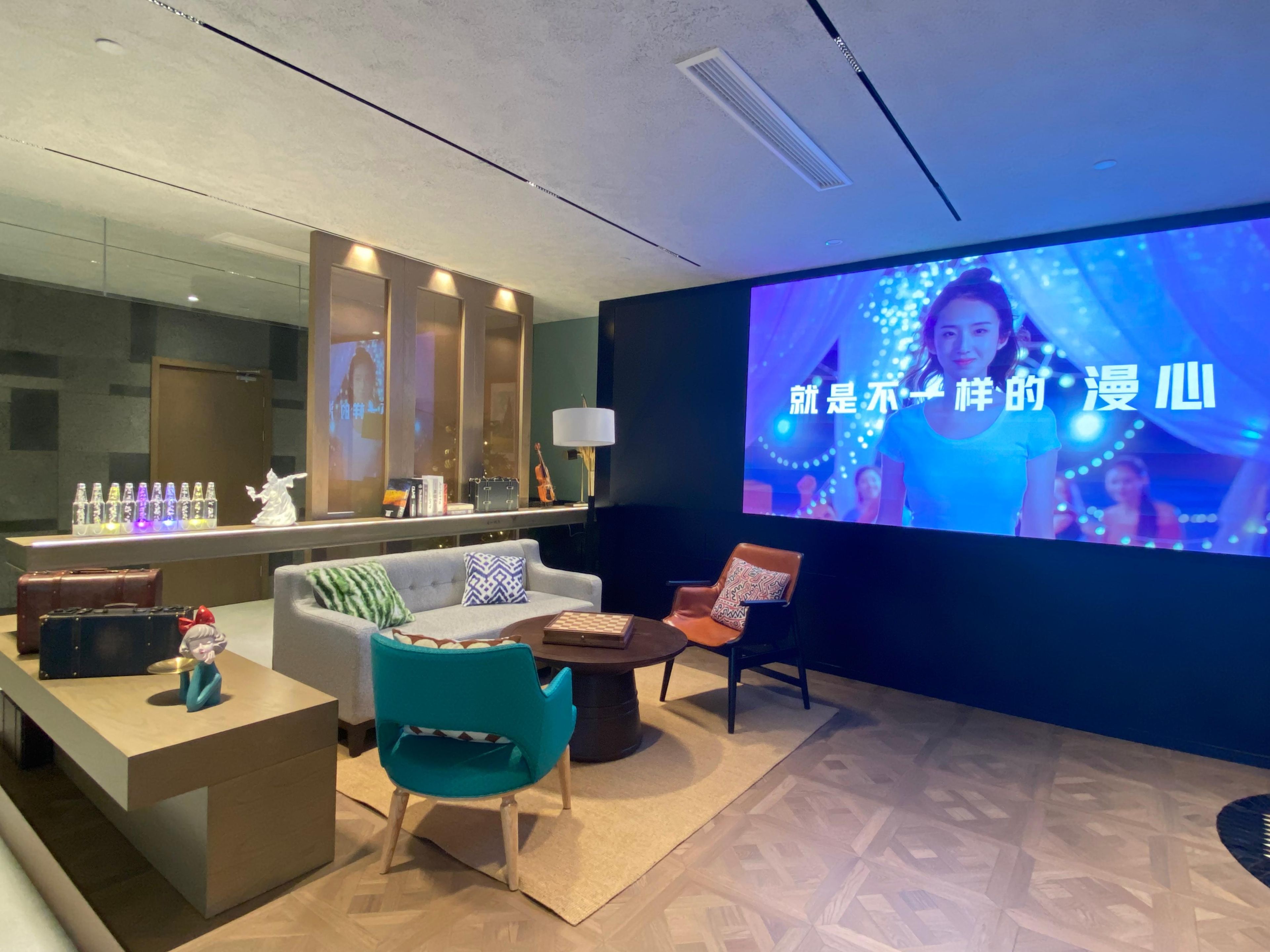 A modern lounge area with a large screen displaying a program, a sofa, a teal chair, a small table with a chessboard, and a bar area with bottles.