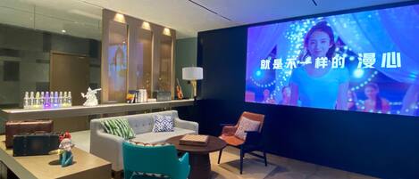 A modern lounge area with a large screen displaying a program, a sofa, a teal chair, a small table with a chessboard, and a bar area with bottles.
