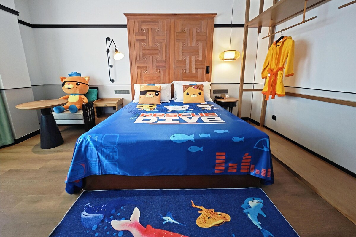 Un dormitorio con cama, mesita de noche, cómoda, silla, mesa y un perchero con toallas.