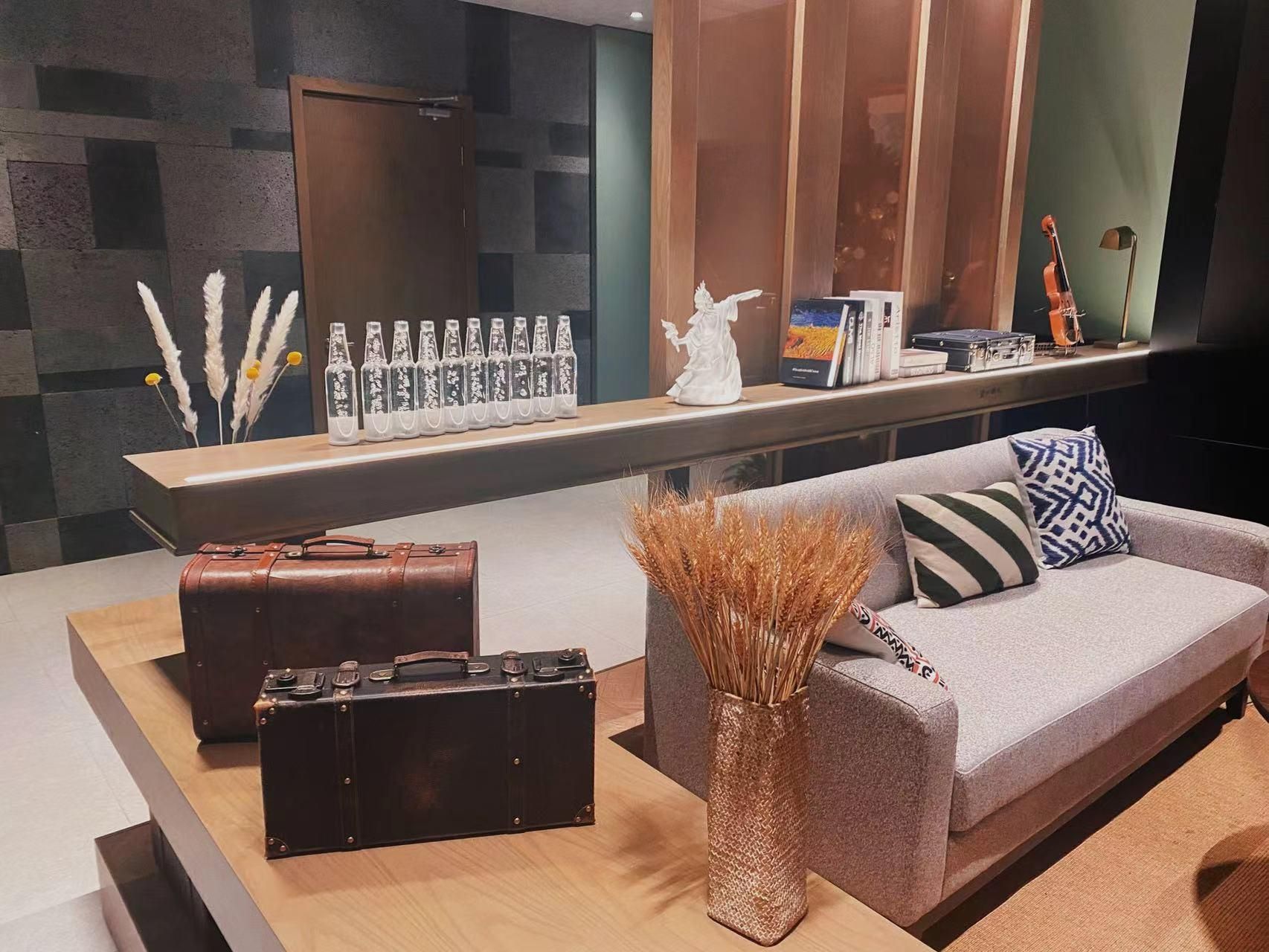A modern living area with a grey sofa, a small table with a vase of dried plants, a brown leather suitcase, and a shelf with decorative items and books.