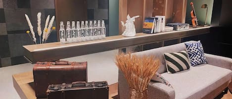 Un espace de vie moderne avec un canapé gris, une petite table sur laquelle est posé un vase contenant des plantes séchées, une valise en cuir marron et une étagère avec des objets décoratifs et des livres.
