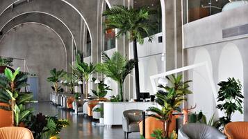 A modern hotel lobby with curved ceiling arches, a reception desk, seating areas, and various indoor plants.