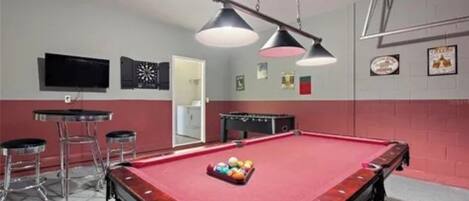 Una mesa de billar con superficie de fieltro rojo, dos taburetes de bar, un televisor montado en la pared y una lámpara en el techo.