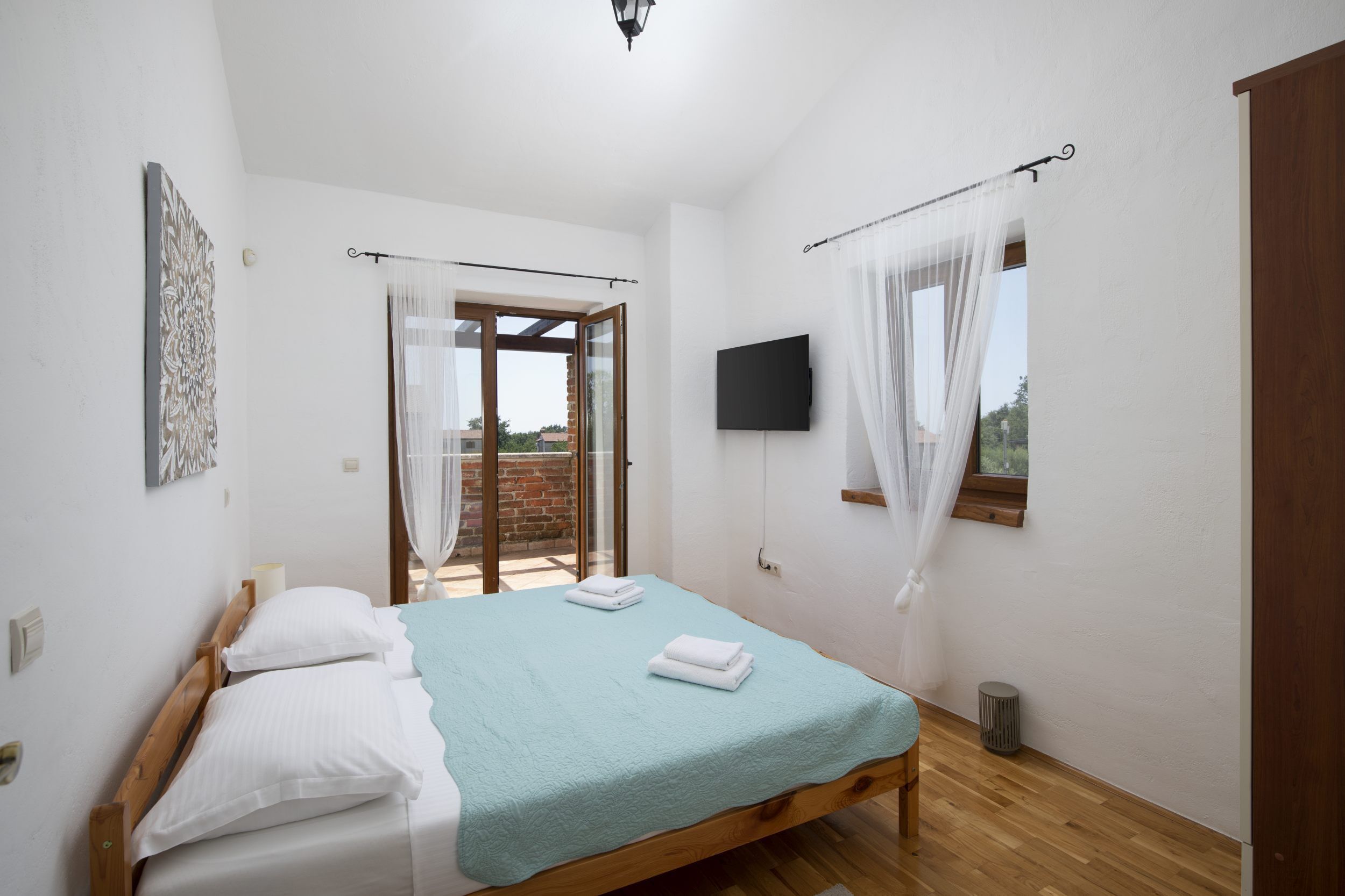Ein Schlafzimmer mit einem Bett, einem Fernseher, einem Fenster mit Vorhängen und einer Tür zu einem Außenbereich.