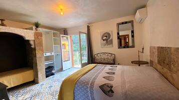 A bedroom with a bed, a chair, a small table, a mirror, and a balcony with a view of greenery.