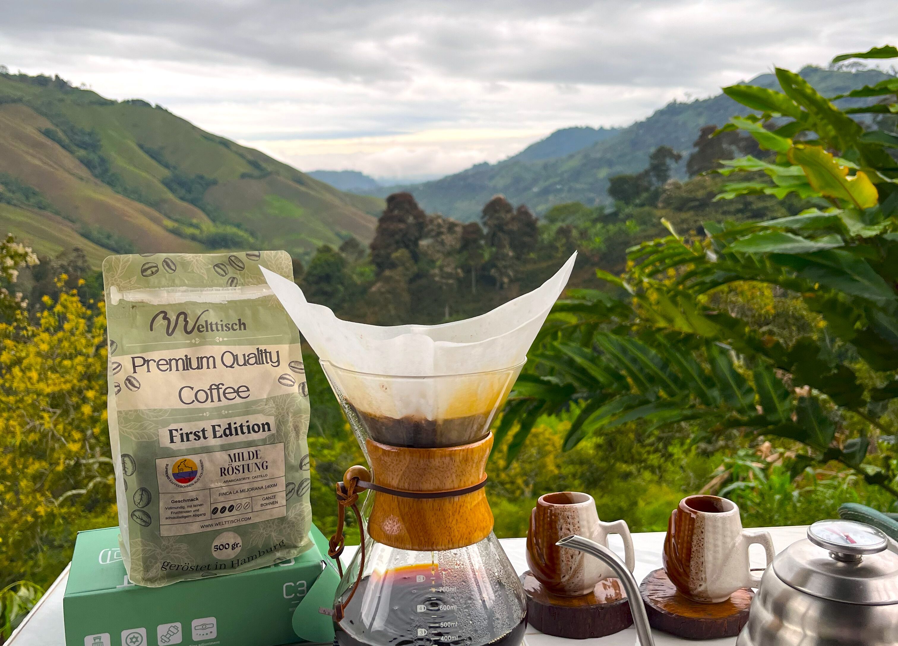 Una cafetera con un paquete de café Weltsch Premium Quality, una taza y una tetera sobre una mesa con vistas panorámicas a colinas y follaje en el fondo.