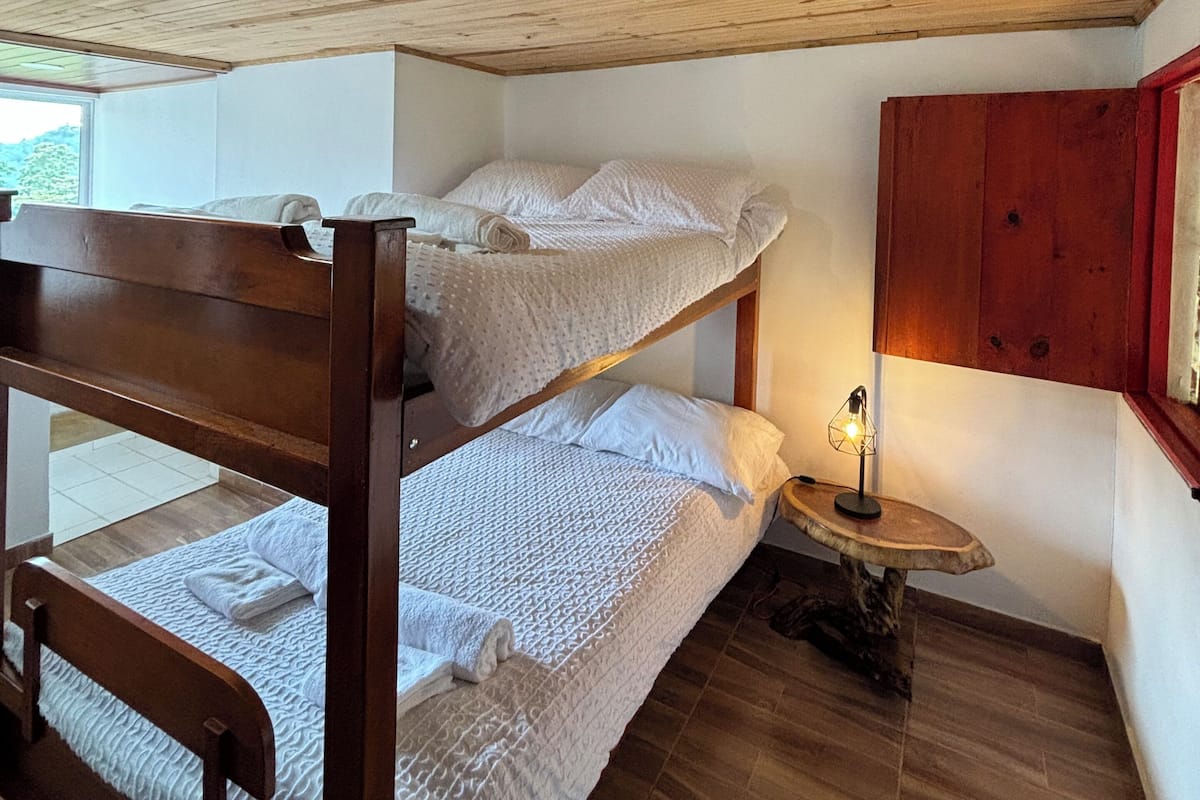 Une chambre avec un lit superposé, un plafond en bois, une petite table ronde avec une lampe et une fenêtre laissant entrer la lumière naturelle.