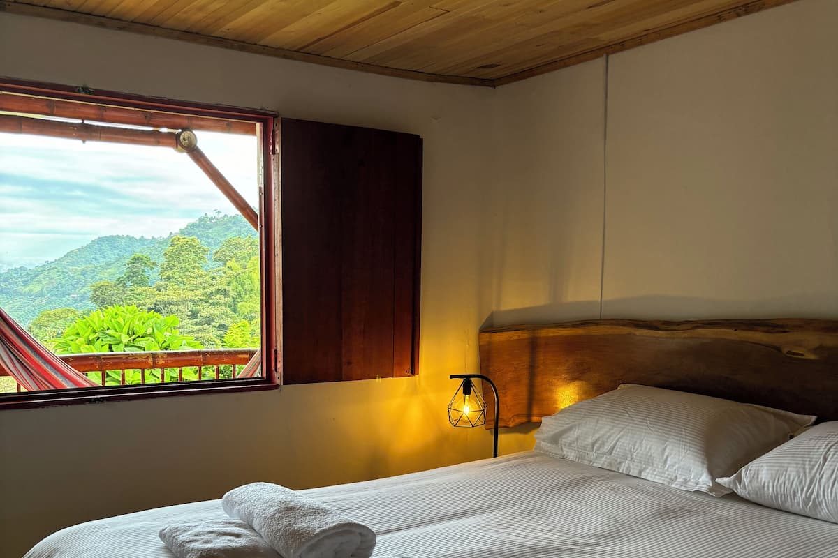 Un lit avec du linge de lit blanc et des serviettes enroulées, une tête de lit en bois, une lampe de chevet et une fenêtre donnant sur un paysage verdoyant et vallonné.