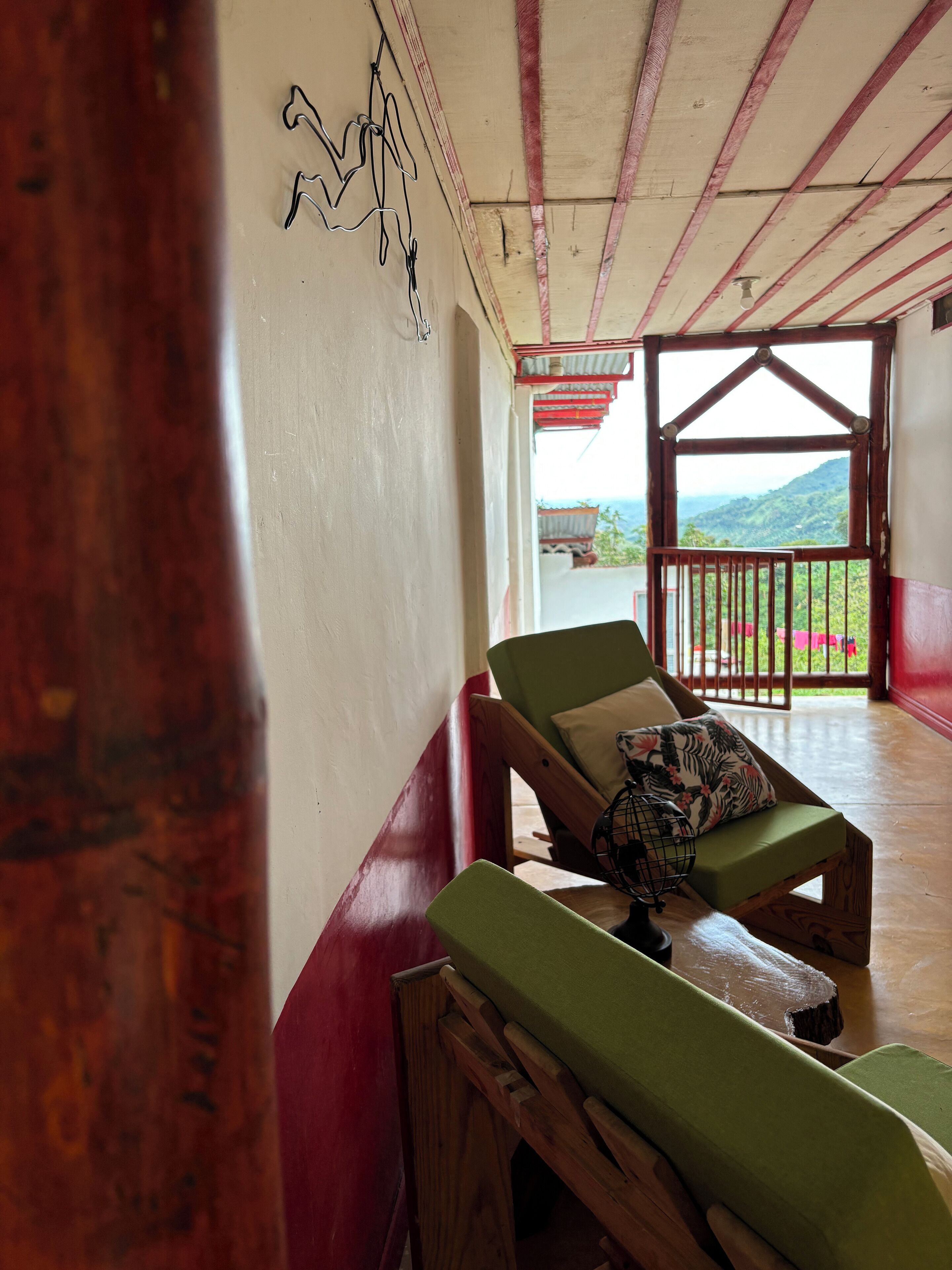 A room with a wooden bench and a green cushioned chair, a wall with a simple line drawing, and a view of a mountainous landscape through an open doorway.