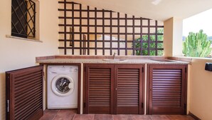 A compact laundry area with a washing machine, wooden cabinets, and a window with a view of greenery. - Casa Marebl in Palau (Palau)