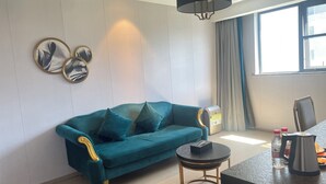 Un sofá color turquesa con detalles dorados, una mesita de centro redonda, una lámpara negra y una ventana con cortinas.