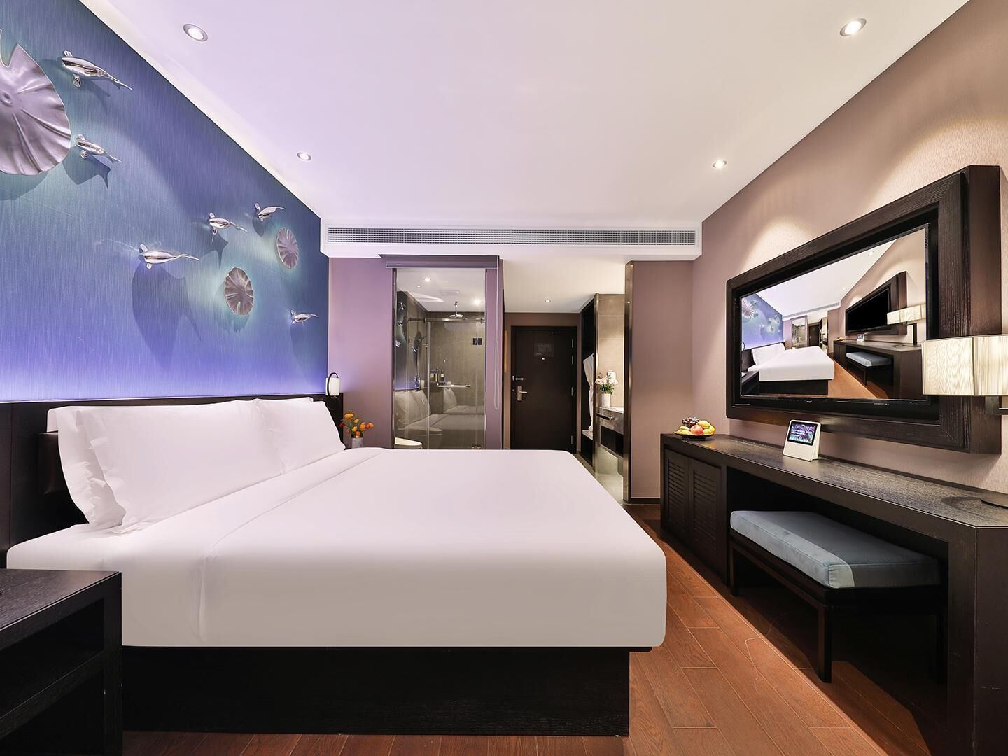Una habitación de hotel moderna con una cama grande, un televisor de pantalla plana, un escritorio y un baño visible a través de una puerta de vidrio.
