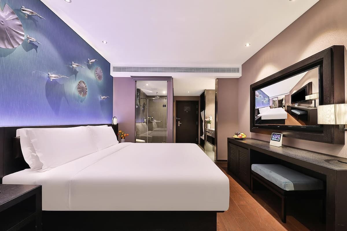 A modern hotel room with a large bed, a flat-screen TV, a desk, and a bathroom visible through a glass door.