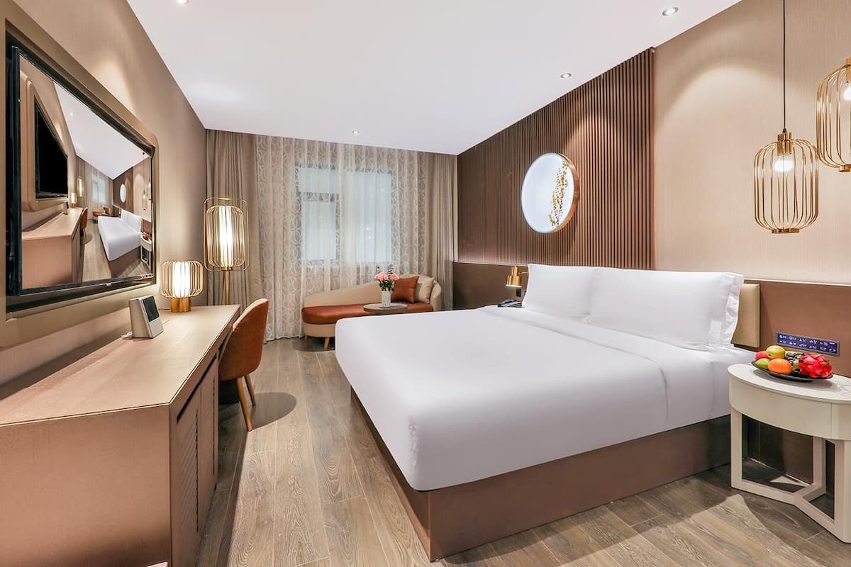 A modern hotel room with a large bed, a desk, a chair, a round mirror, and a bedside table with fruit.