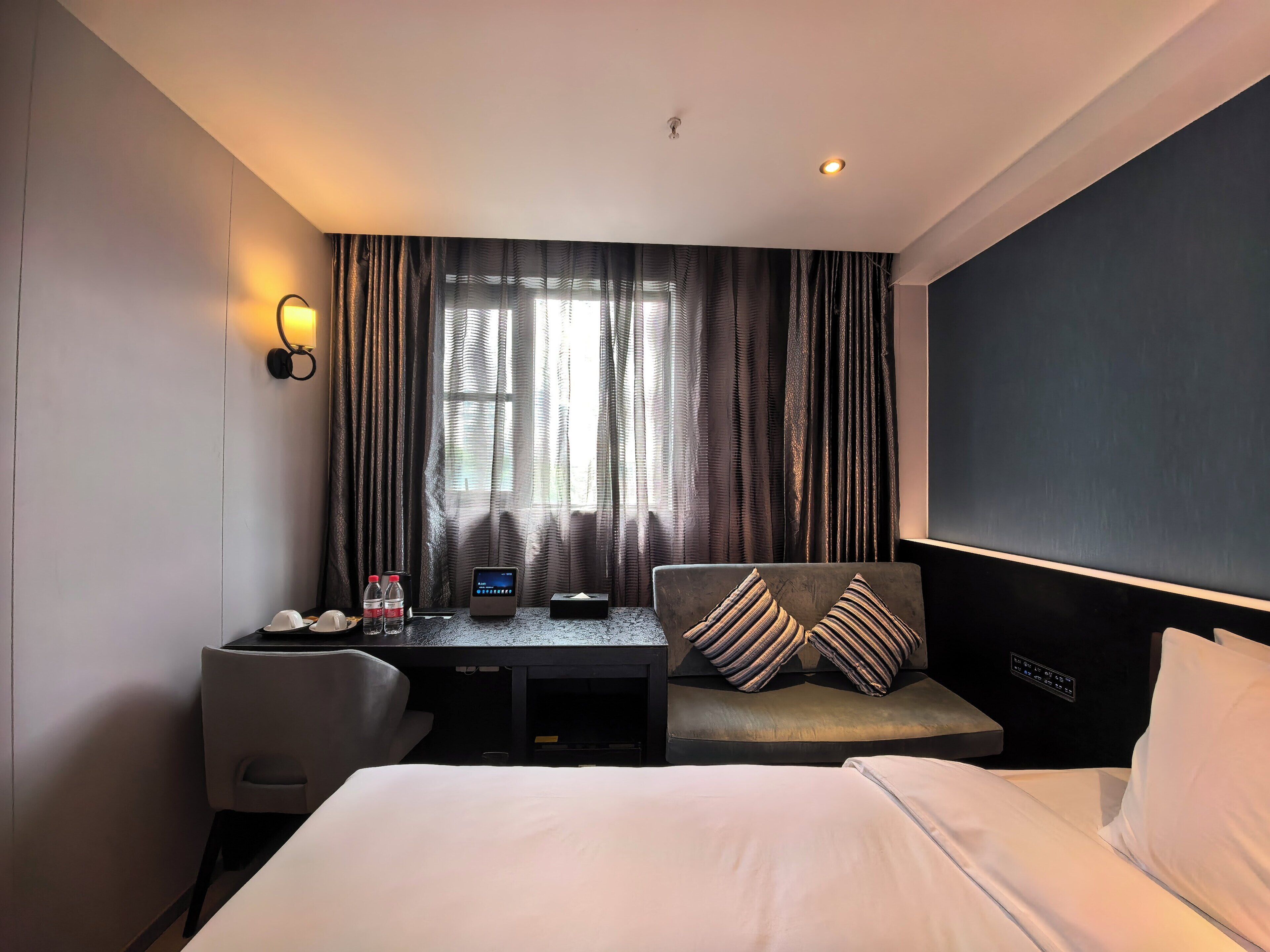 Une chambre d’hôtel comprenant un lit, un bureau, une chaise, une fenêtre avec des rideaux et un panneau de commande fixé au mur.