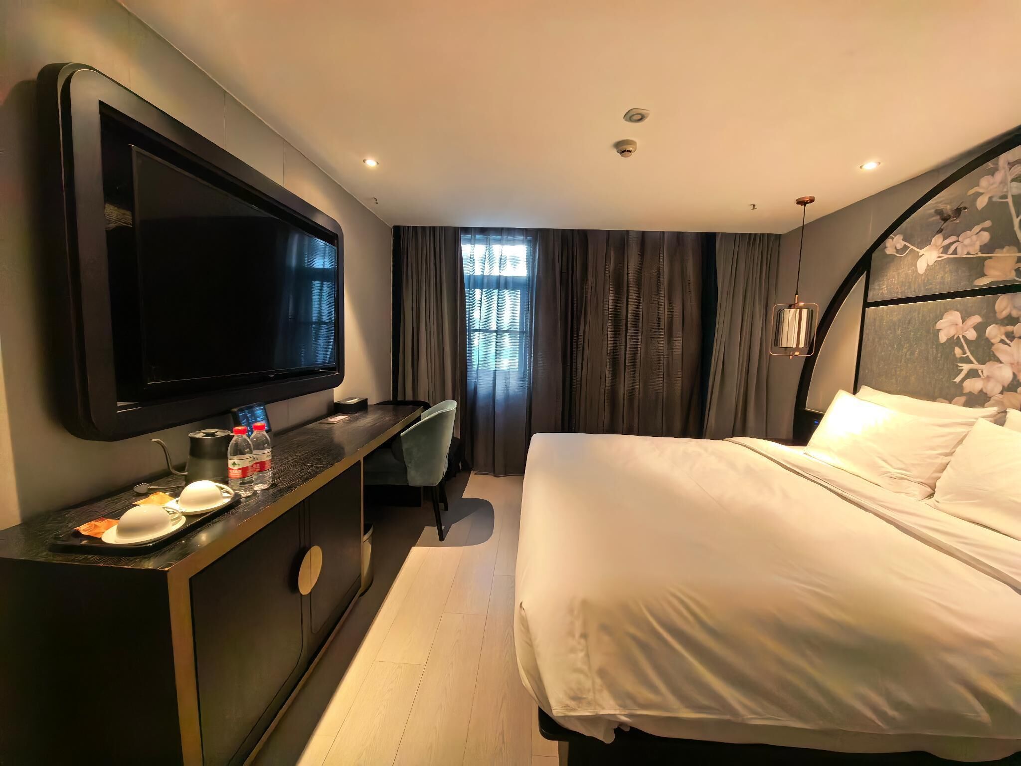 A hotel room with a bed, a desk with a flat-screen TV, a minibar, and a window with curtains.