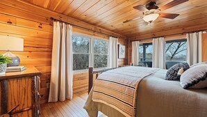Une chambre confortable aménagée dans un chalet en bois, avec un lit, un bureau doté d’une lampe et de grandes fenêtres donnant sur la forêt.
