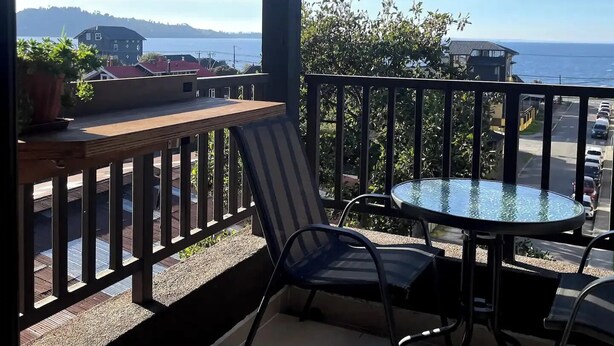 A balcony with a table and chairs, overlooking a coastal view with buildings and a road. - Beautiful Apartment With Terrace and Lake View (Puerto Varas)