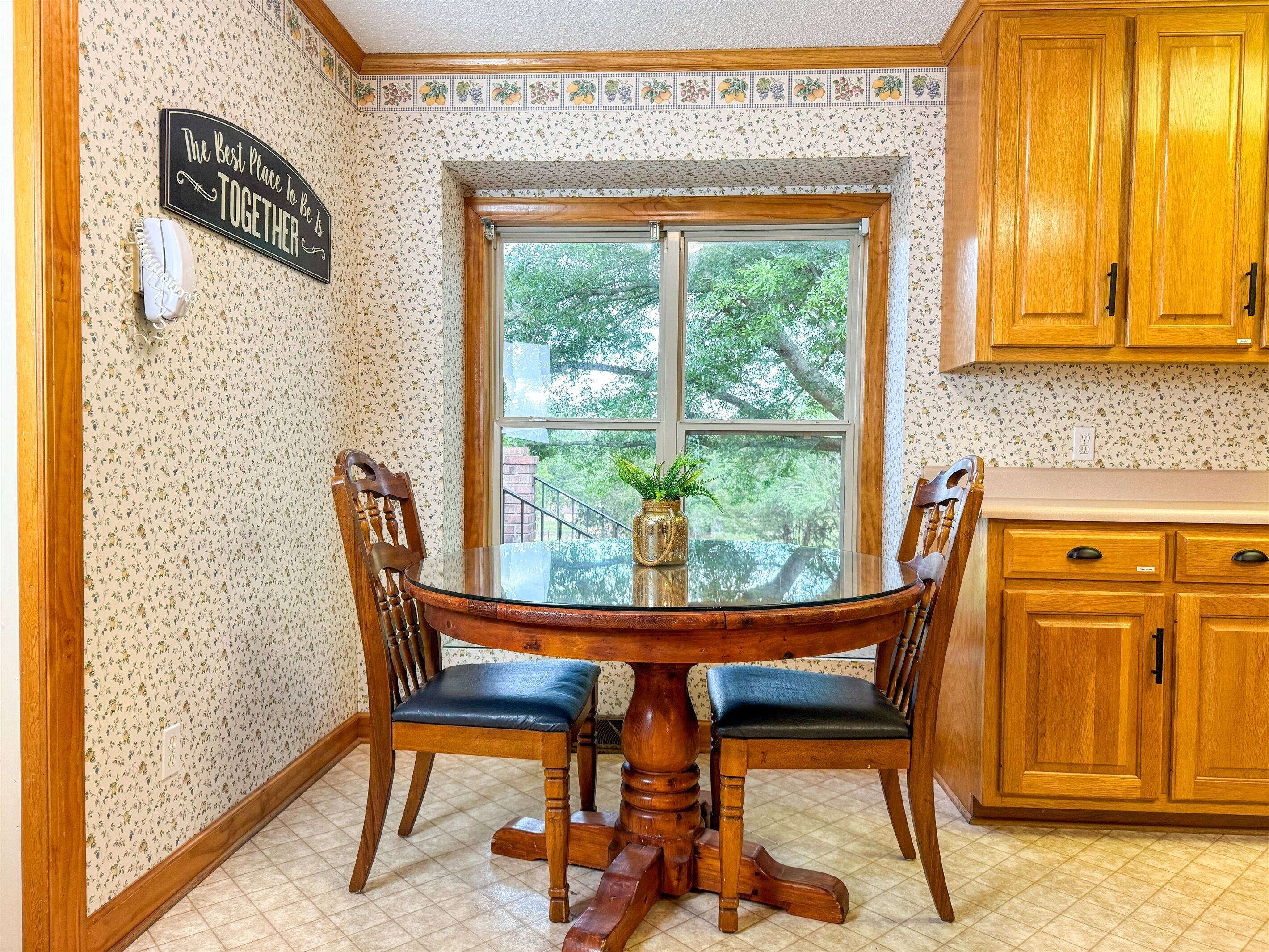 A kitchen with a round wooden table, two chairs, a window with a view of trees, and wooden cabinets.